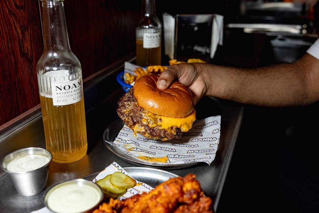 alguien con una hamburguesa en la mano junto a una cerveza y un plato de alitas