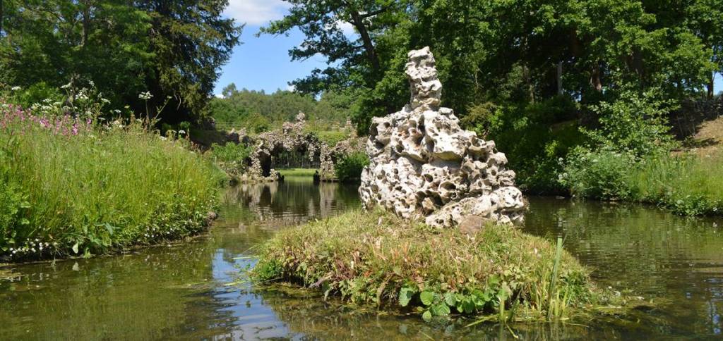 ein Steinhaufen auf einer Rasenfläche mitten in einem See, in der Nähe der Kristallgrotte im Painshill Park
