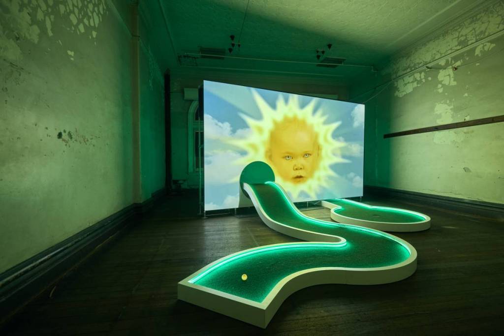 um campo de minigolfe iluminado com um ecrã gigante no meio a mostrar o sol dos Teletubbies