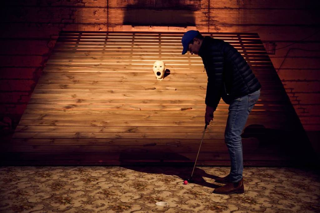 alguém a preparar-se para dar uma tacada numa bola de golfe em direção a uma cabeça a gritar no meio de uma rampa num campo de minigolfe, com a boca bem aberta à espera da bola