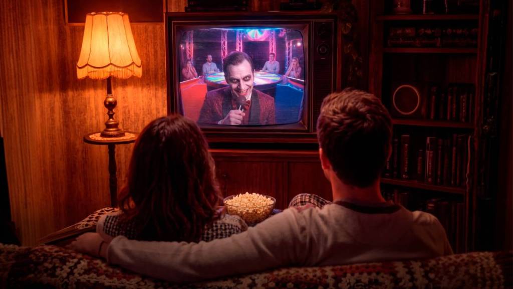Zwei Personen sind von hinten zu sehen, wie sie auf einem Sofa sitzen und einem gruseligen Clown zusehen, der auf einem Bildschirm auf sie zeigt, dazwischen liegt Popcorn