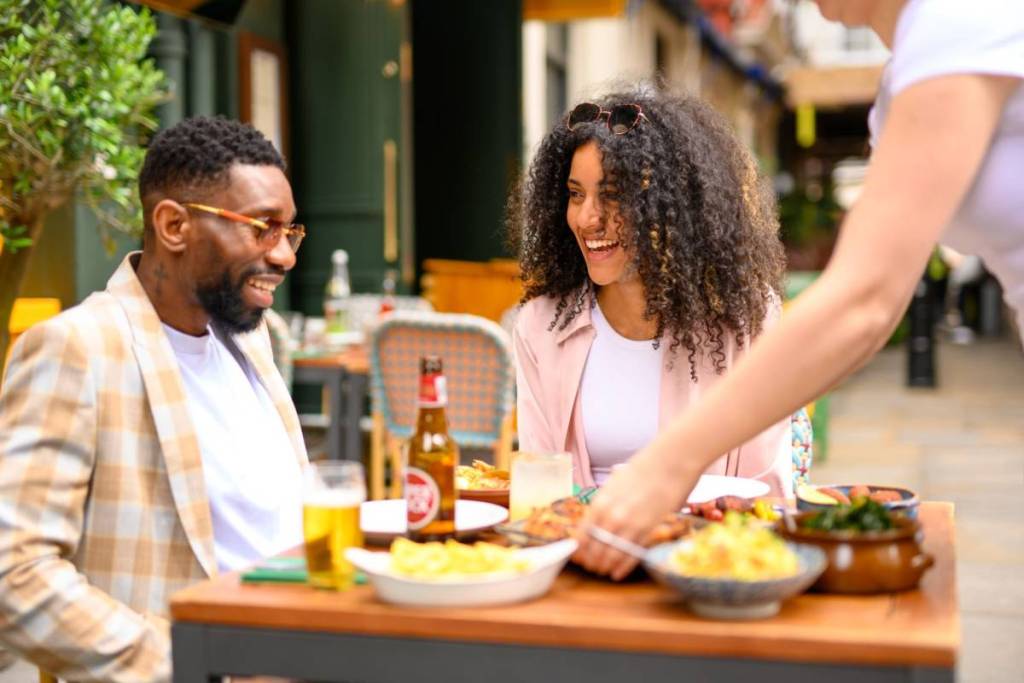 deux personnes assises à une table à l’extérieur d’un restaurant tandis qu’un serveur leur sert leur repas