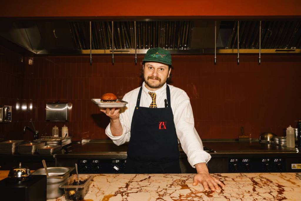 un chef en tablier, coiffé d’une casquette et vêtu d’une chemise et d’une cravate, tenant une assiette sur laquelle trône un burger en édition limitée