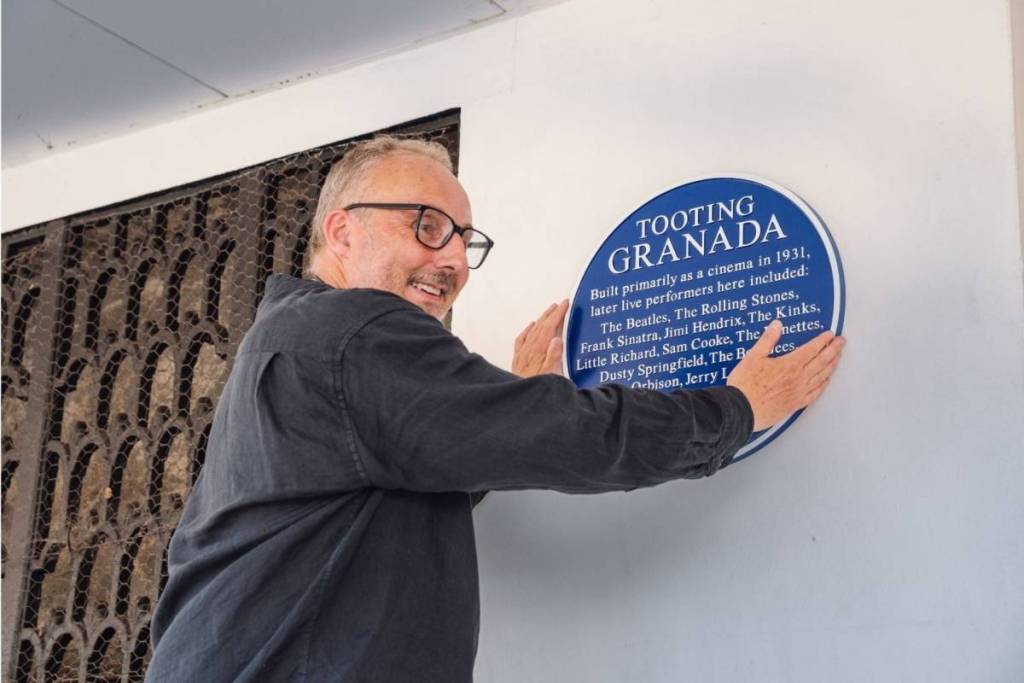 A man smiles as he hangs a blue plaque on a white wall, thw words "Tooting Granada" is visible. 