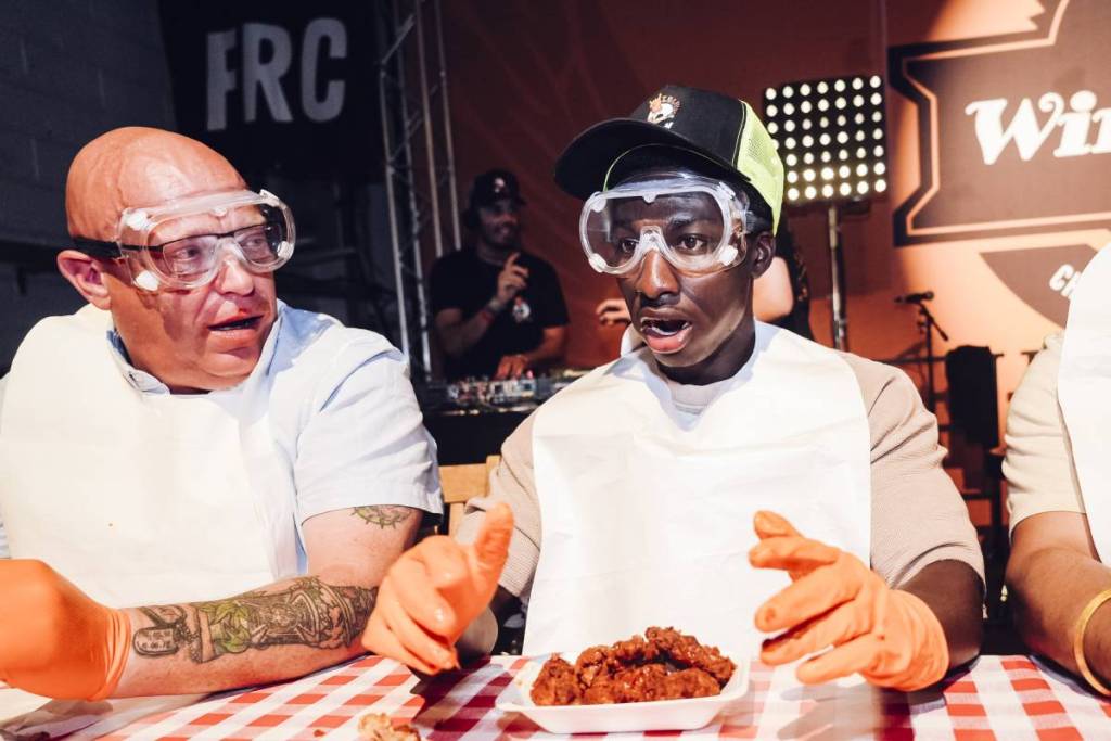 personas con mascarillas y guantes de seguridad en un concurso de comer alitas, que se quedan boquiabiertas por el picante de las alitas