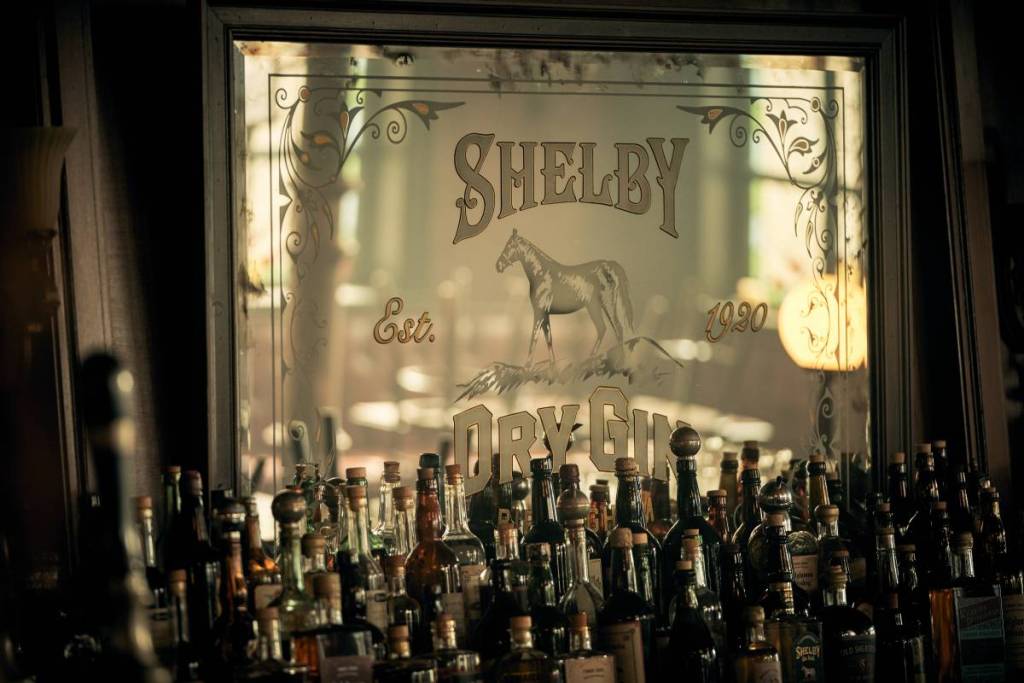 Ein Regal voller Spirituosenflaschen unter einem Spiegel mit der Aufschrift „Shelby Gin“ – sieht aus wie eine Kulisse aus „Peaky Blinders“