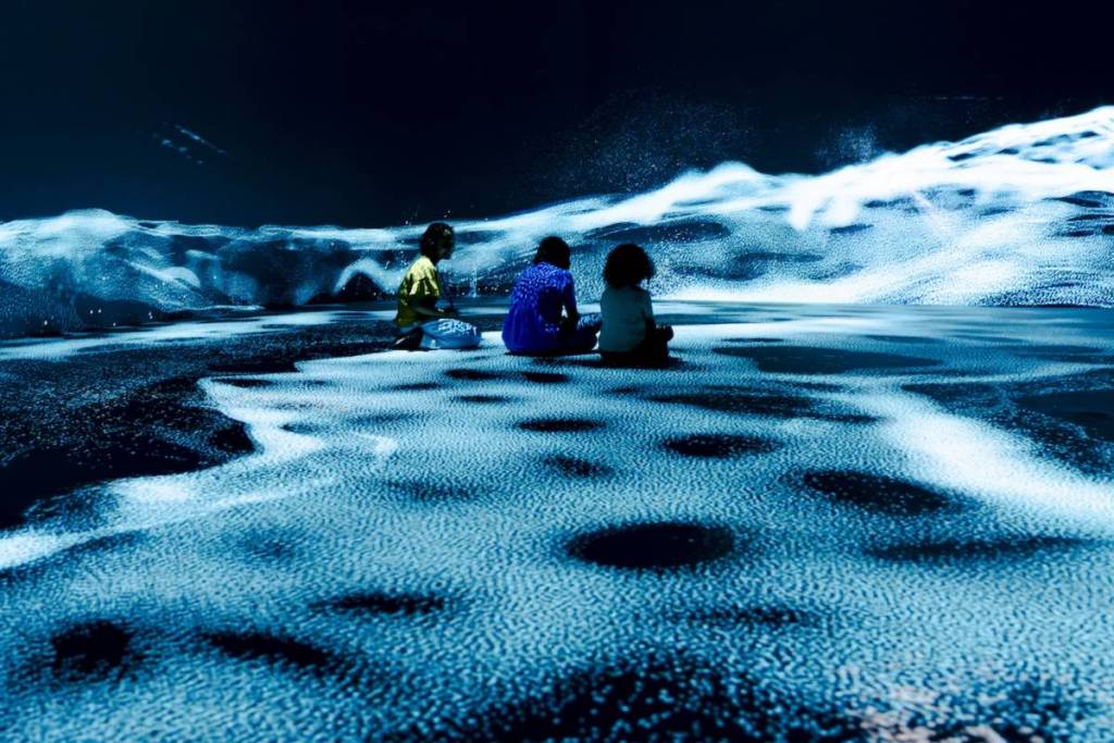 Tres niños están sentados con las piernas cruzadas en el suelo, en una sala inmersiva con proyecciones abstractas de lo que parecen dunas de arena azules brillantes