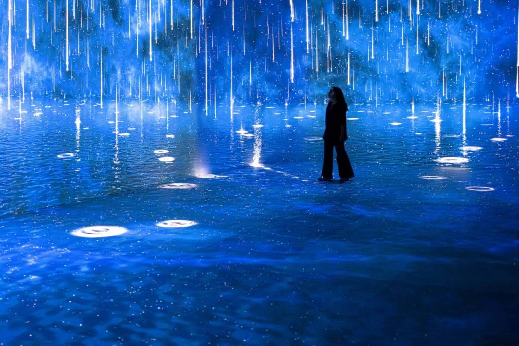 una figura solitaria recorre una sala inmersiva con proyecciones de lluvia dorada cayendo sobre aguas azul oscuro
