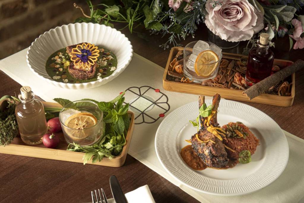 Ein Tisch voller besonderer Cocktails, umgeben von Kräutern und Flaschen mit Zutaten, sowie zwei von der Flora inspirierte Gerichte der indischen Küche