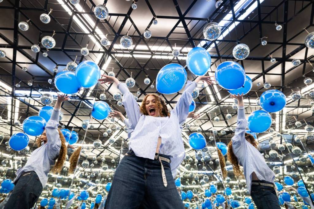 a joyful looking person throwing their hands up in a mirrored room full of suspended balloons and with reflections dotted around them