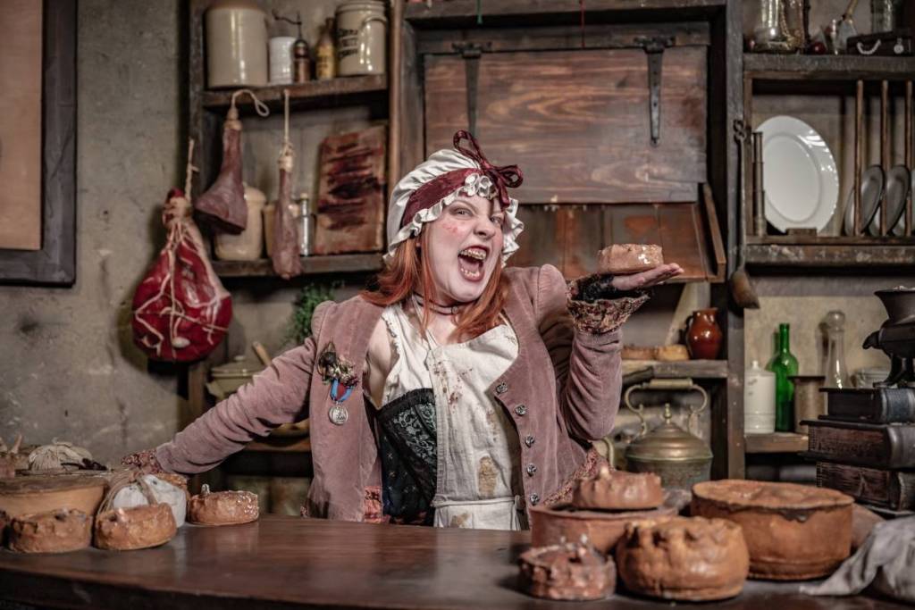 hält eine als Mrs. Lovett verkleidete Schauspielerin eine Pastete hoch und scheint in die Kamera zu kichern