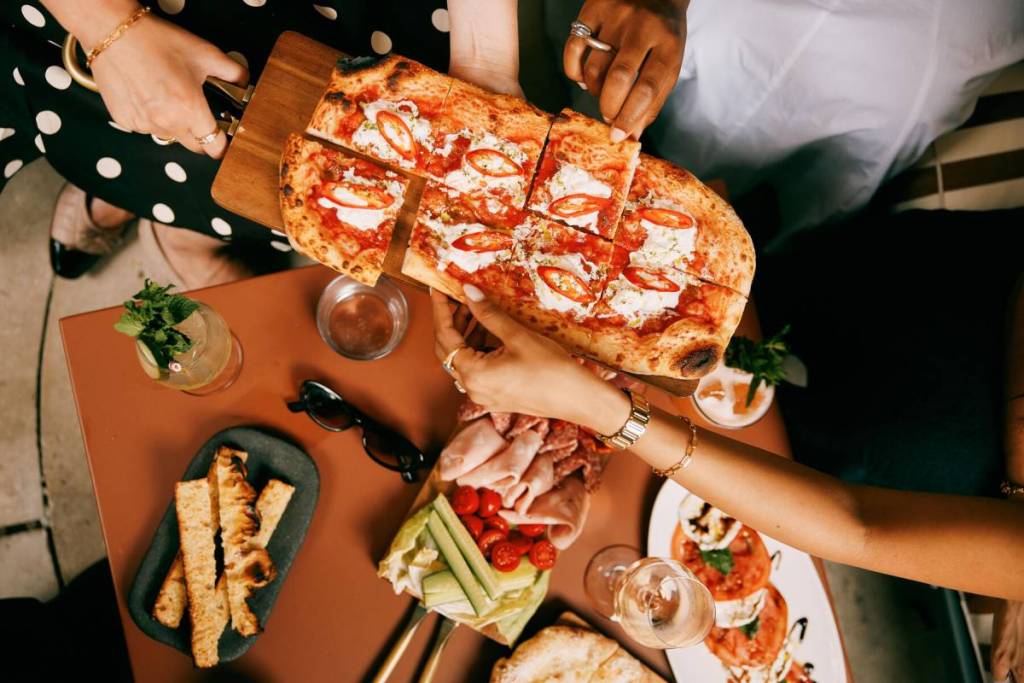mensen die stukjes pizza pakken van een aangeboden bord, dat boven de bordjes met hapjes en voorgerechten wordt gehouden