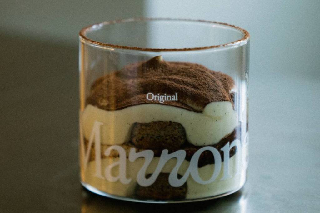 Un tarro de cristal sobre una mesa con un cremoso tiramisú dentro y una capa de chocolate marrón por encima