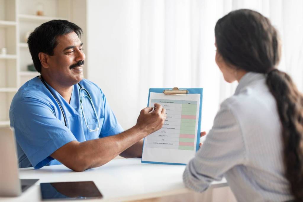 Médico indiano a mostrar uma prancheta com um formulário de exames médicos a uma paciente numa clínica; terapeuta masculino de uniforme a fazer perguntas a uma mulher sorridente durante um check-up do NHS num hospital; Espaço livre