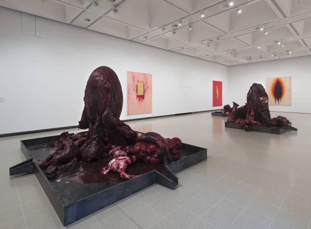 een zaal op de Anish Kapoor-tentoonstelling in de Hayward Gallery met schilderijen aan de muren en sculpturale werken midden in de zaal, die door suggestieve kleuren, vormen en combinaties gruwelijke, bloederige taferelen lijken uit te beelden
