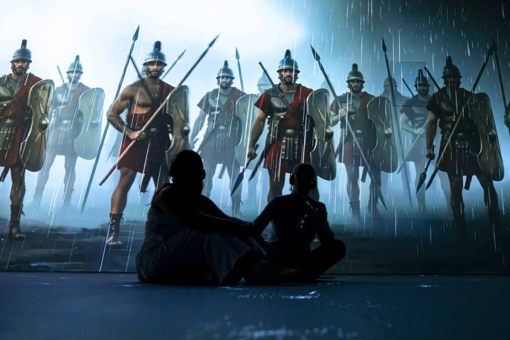 Deux personnes sont assises par terre à l’exposition immersive Timewalk à Londres, se découpant en silhouette sur un fond de soldats romains de l’Antiquité