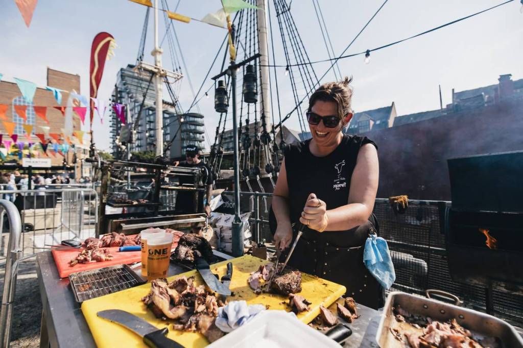 a food vendor at meatopia, stood at a stand with a large boat behind them, carving meat to portion into servings for festival attendees