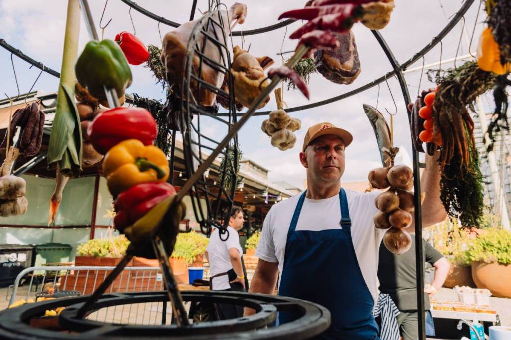 a cook tending to an array of hanging vegetables and meats, which are being cooked suspended over a live fire