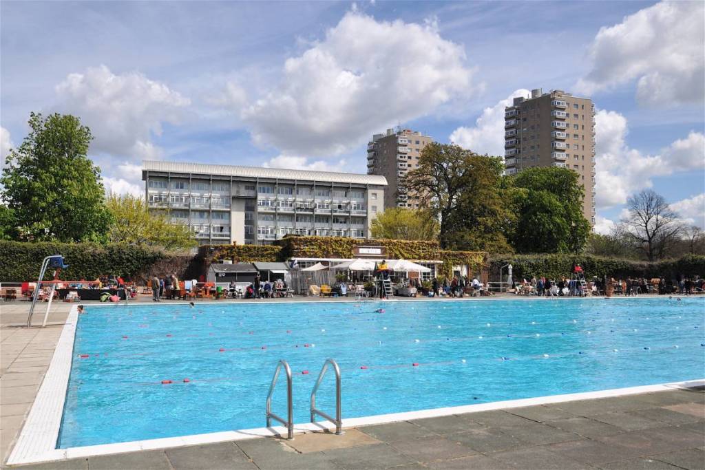 una piscina al aire libre de un azul intenso, rodeada de puestos y casetas donde se vende arte, con grandes bloques de pisos como telón de fondo