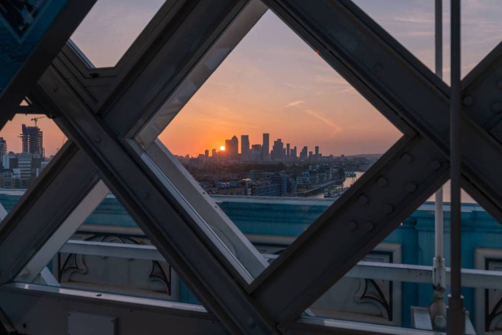 De gebouwen van Canary Wharf bij zonsopgang, gezien door een ruitvormig raam op de glazen loopbrug van de Tower Bridge