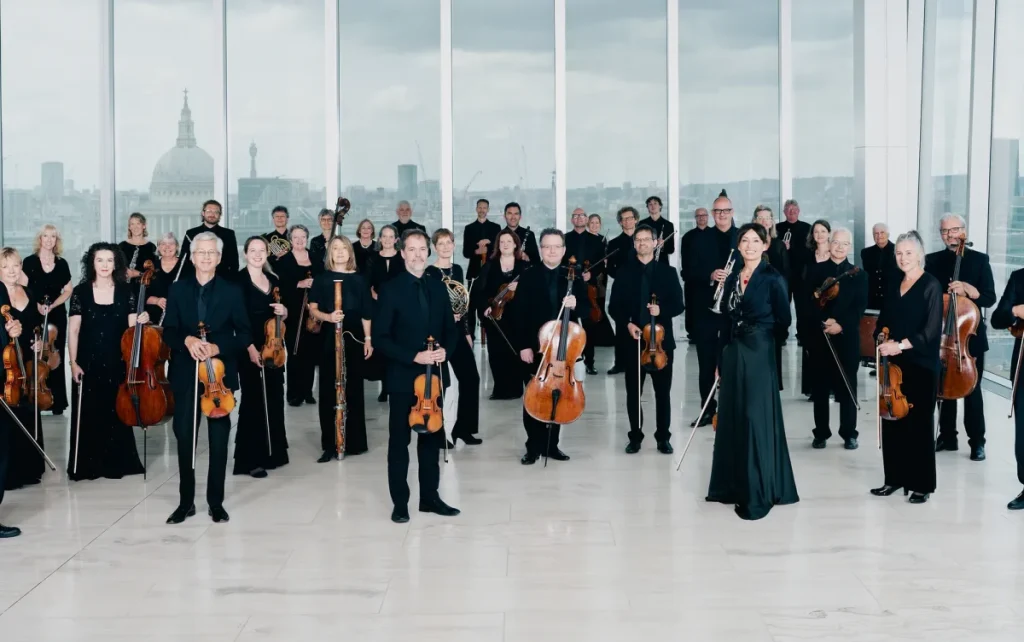 i membri di un’orchestra in piedi in una grande sala con le finestre, con gli strumenti in mano