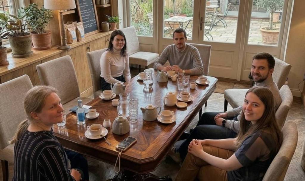 A group of five people sitting around a cafe table with many coffee mugs, looking up at the camera for a group photo