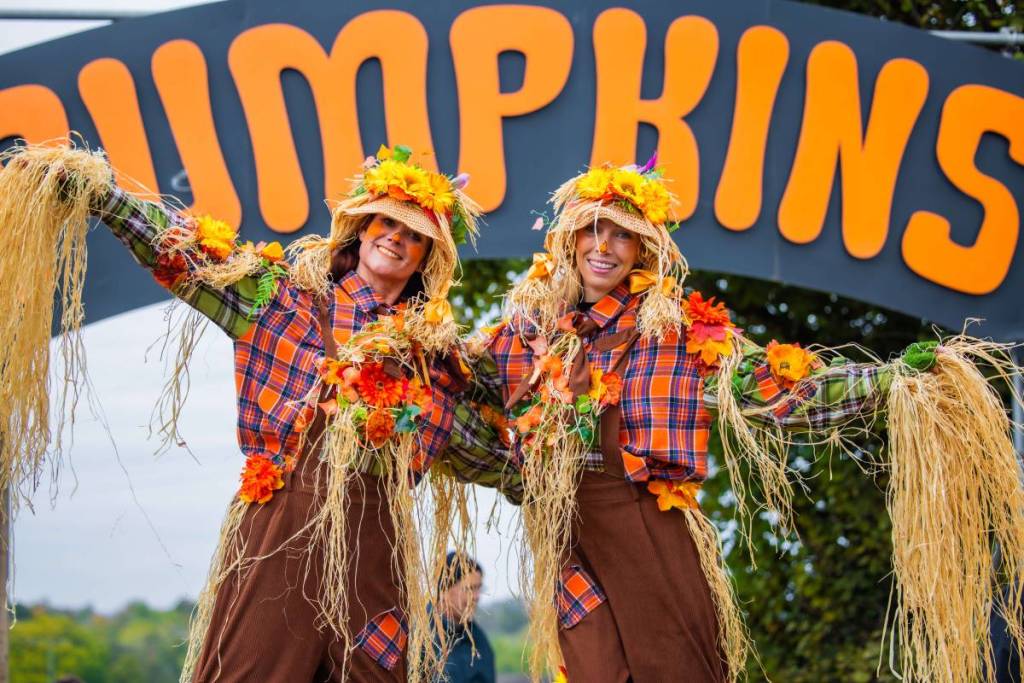 due persone travestite da spaventapasseri a tema zucca davanti a un grande cartello con la scritta PUMPKINS