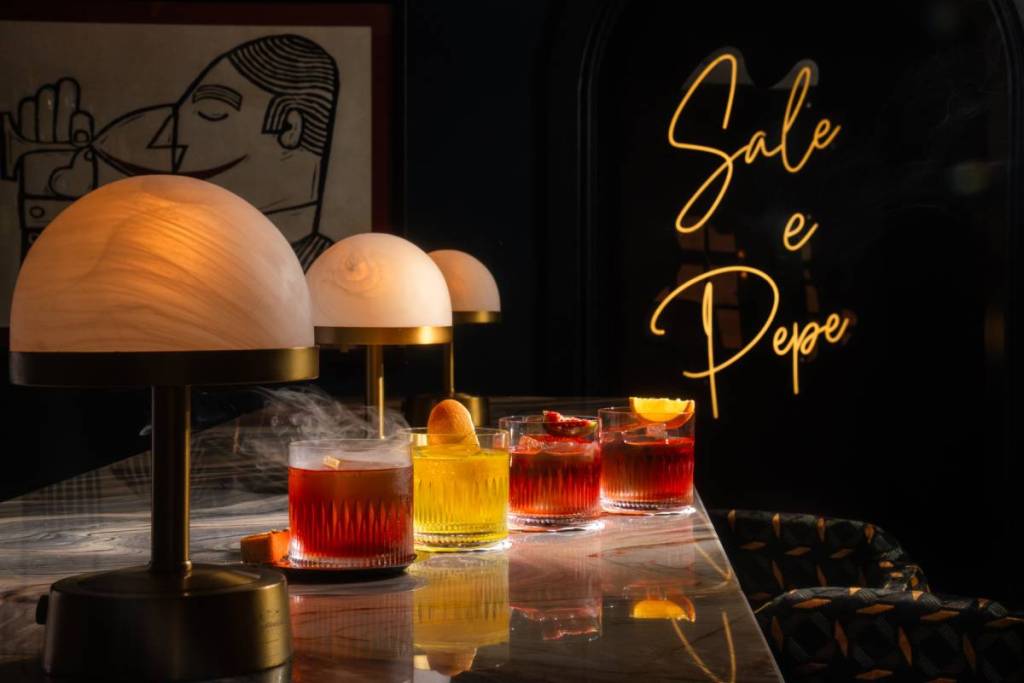 una selezione di Negroni dai colori diversi serviti in un bar sofisticato, accanto a lampade dalla luce soffusa, con un’insegna sullo sfondo che recita “Sale e Pepe” in corsivo