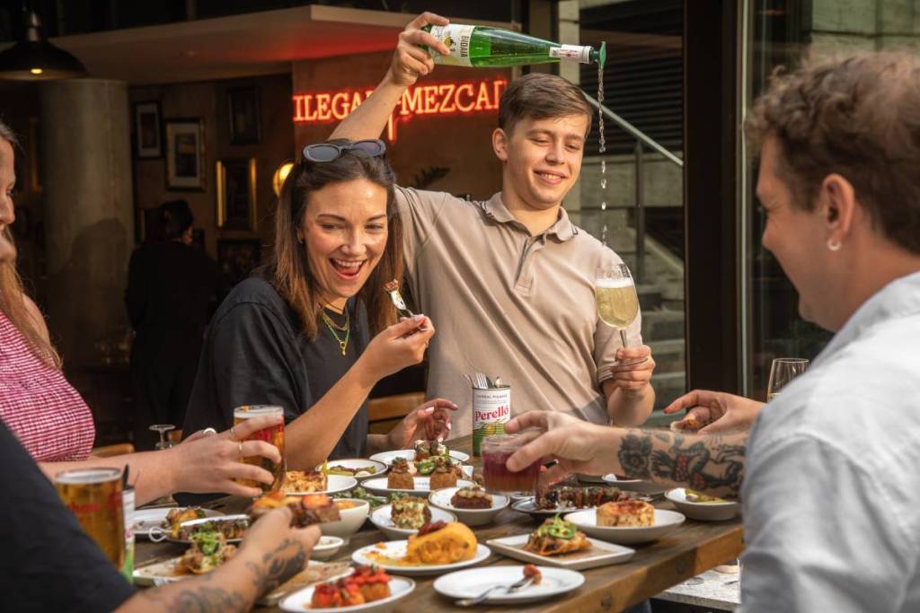 la gente era in piedi o seduta attorno a un tavolo a gustare una vasta selezione di street food basco e tapas, con qualcuno in piedi dietro al tavolo che versava un drink dall’alto da una bottiglia