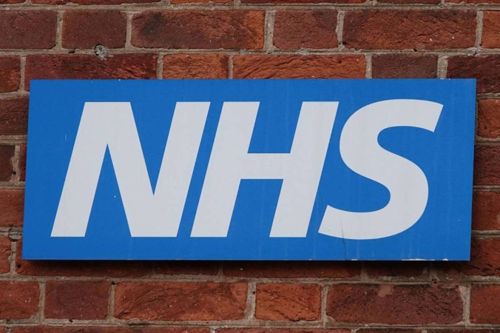 Un panneau bleu et blanc du NHS avec un mur de briques visible en arrière-plan.