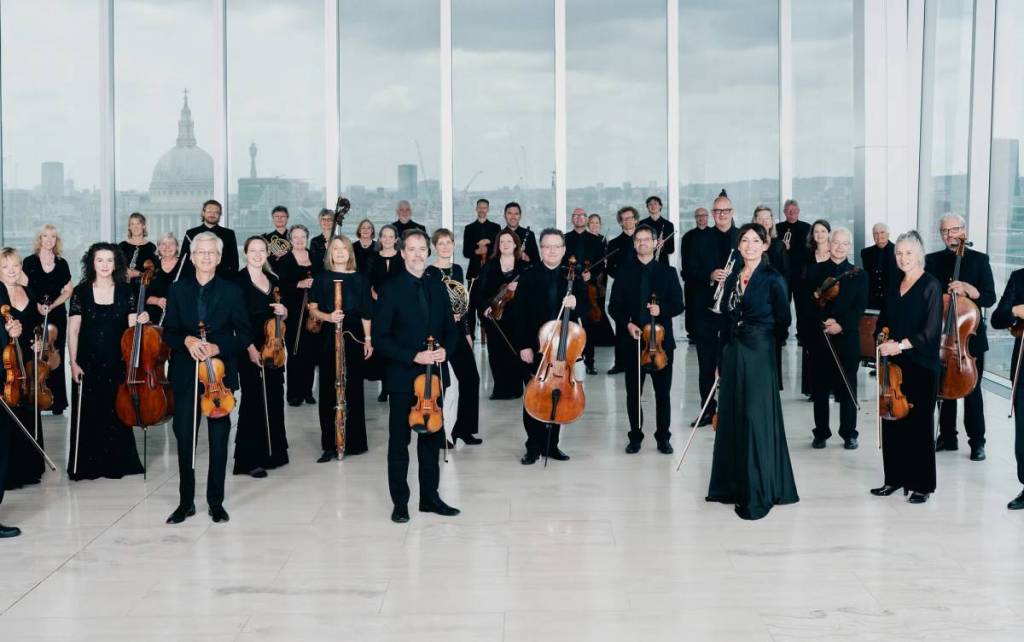 the members of an orchestra standing in a large windowed room, holding their instruments