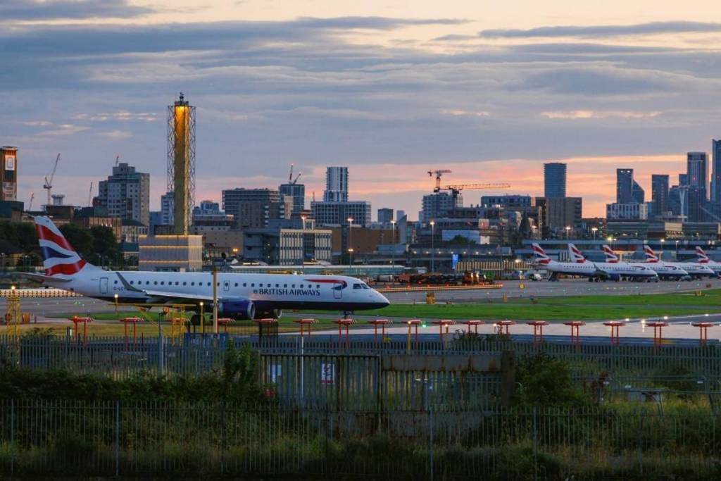 Un aeroporto al tramonto: si vede un aereo della British Airways con altri sullo sfondo, oltre allo skyline di Londra.