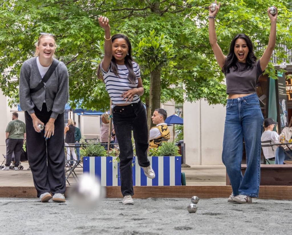 three young women play a game of petanque in king's cross, the woman in the centre throws a ball towards the camera 