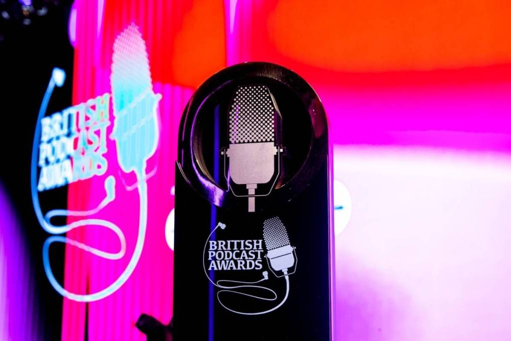 Un premio nero con un microfono da podcast raffigurato, davanti a uno schermo rosso/rosa con una scritta sfocata