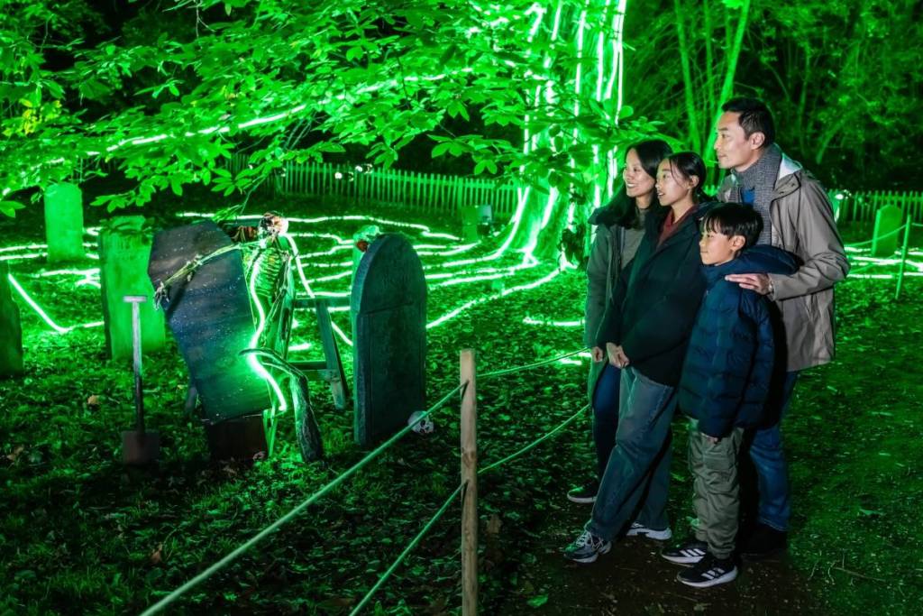 Uma família observa um cenário que representa um cemitério com esqueletos falsos pendurados nas lápides e uma iluminação neon verde sinistra que desce em faixas pelo tronco de uma árvore