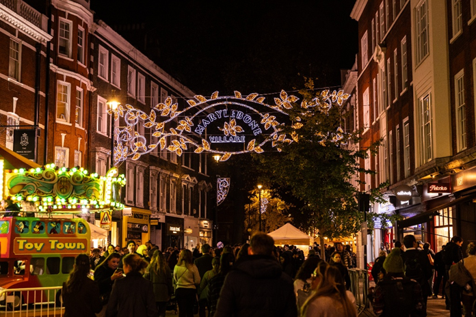 la gente che si aggira in un mercatino invernale serale a tempo determinato, sotto le luci natalizie illuminate con la scritta “Marylebone Village”