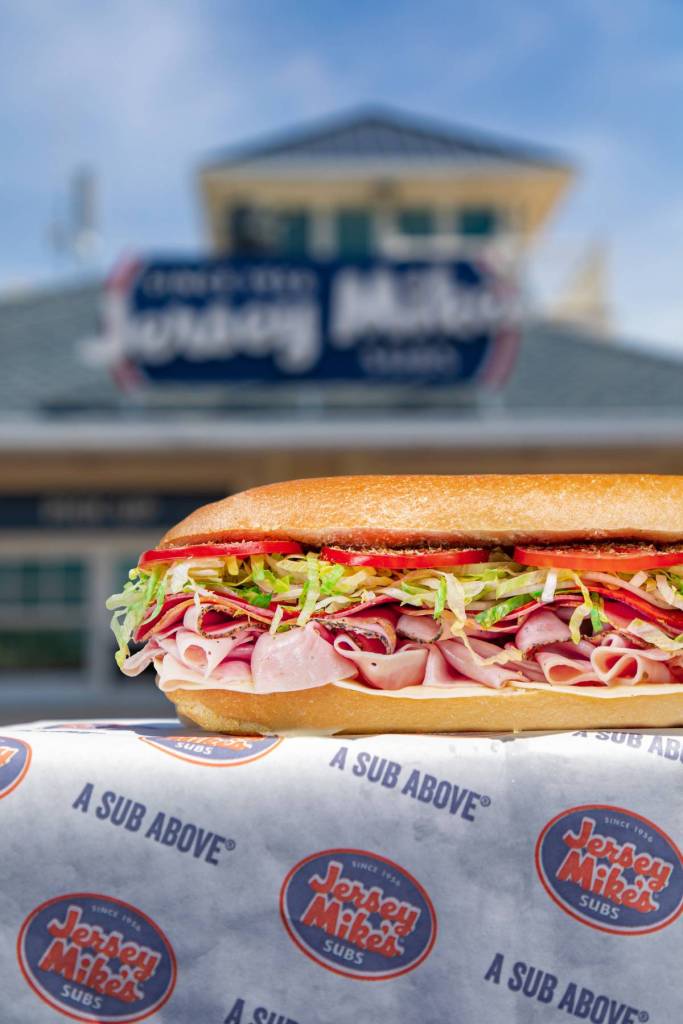 a close-up view of a jersey mike's sub showing plenty of cold cuts, lettuce, and tomatoes, piled high inside a sandwich, against an out-of-focus backdrop of a jersey mike's shop