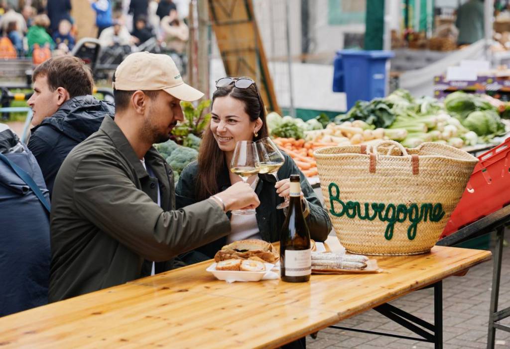 a man and a woman cheersing with two glasses of white wine while sitting at a table in an open air market