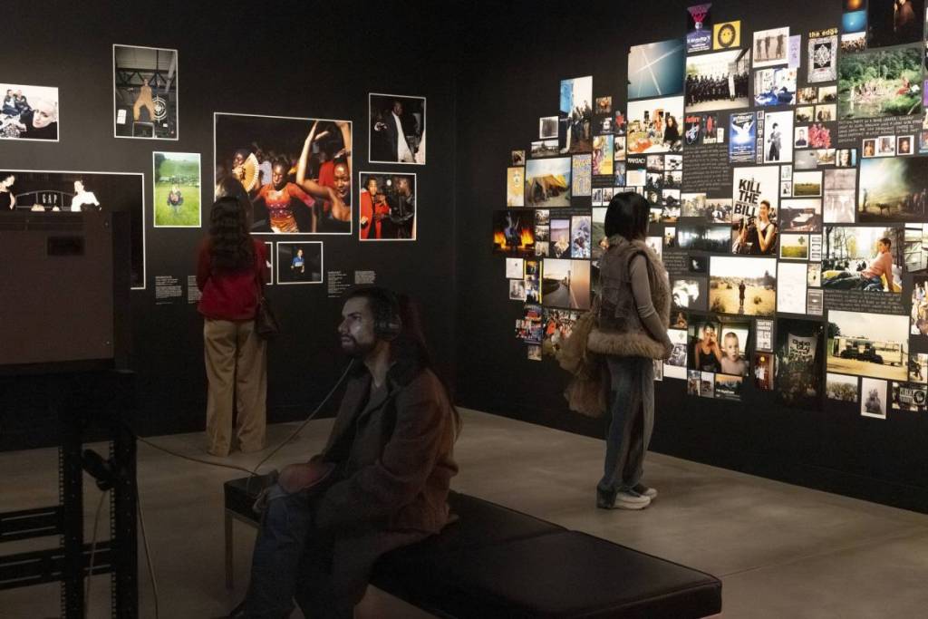 people stood and sat around a dark room looking at walls covered in a mosaic of photos and images of events from the 90s