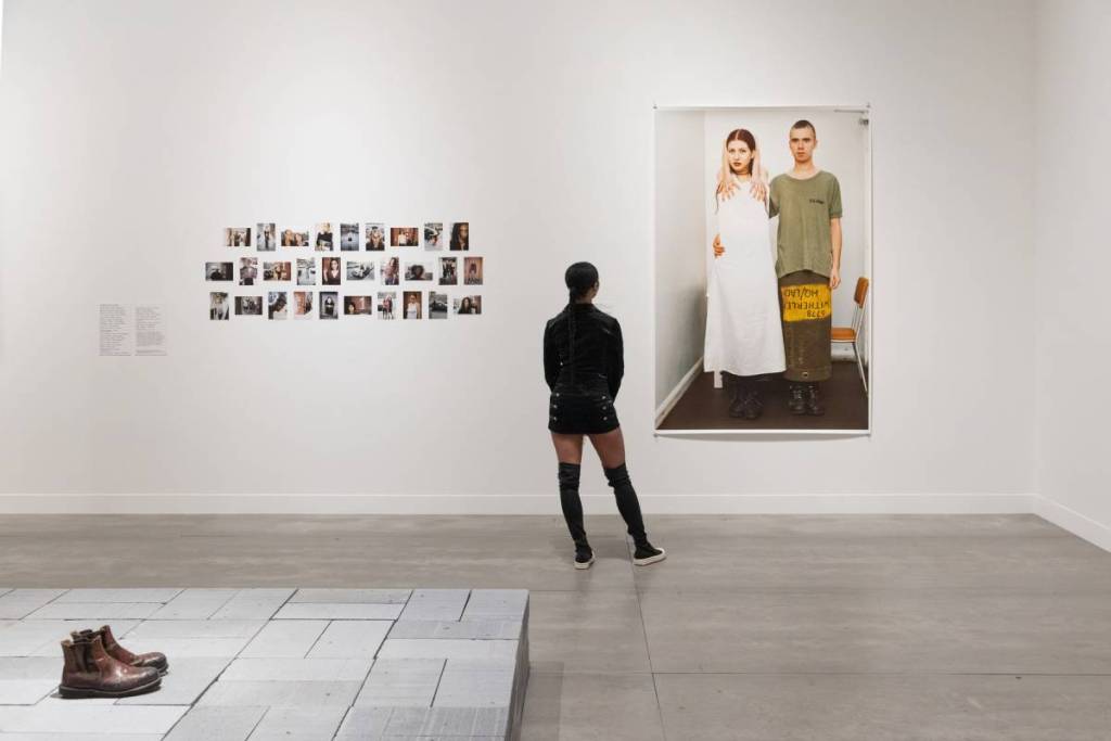 someone standing in a white walled gallery looking at a large photo, next to a collection of tiny photos on the wall. There is a shoe on a platform in the foreground
