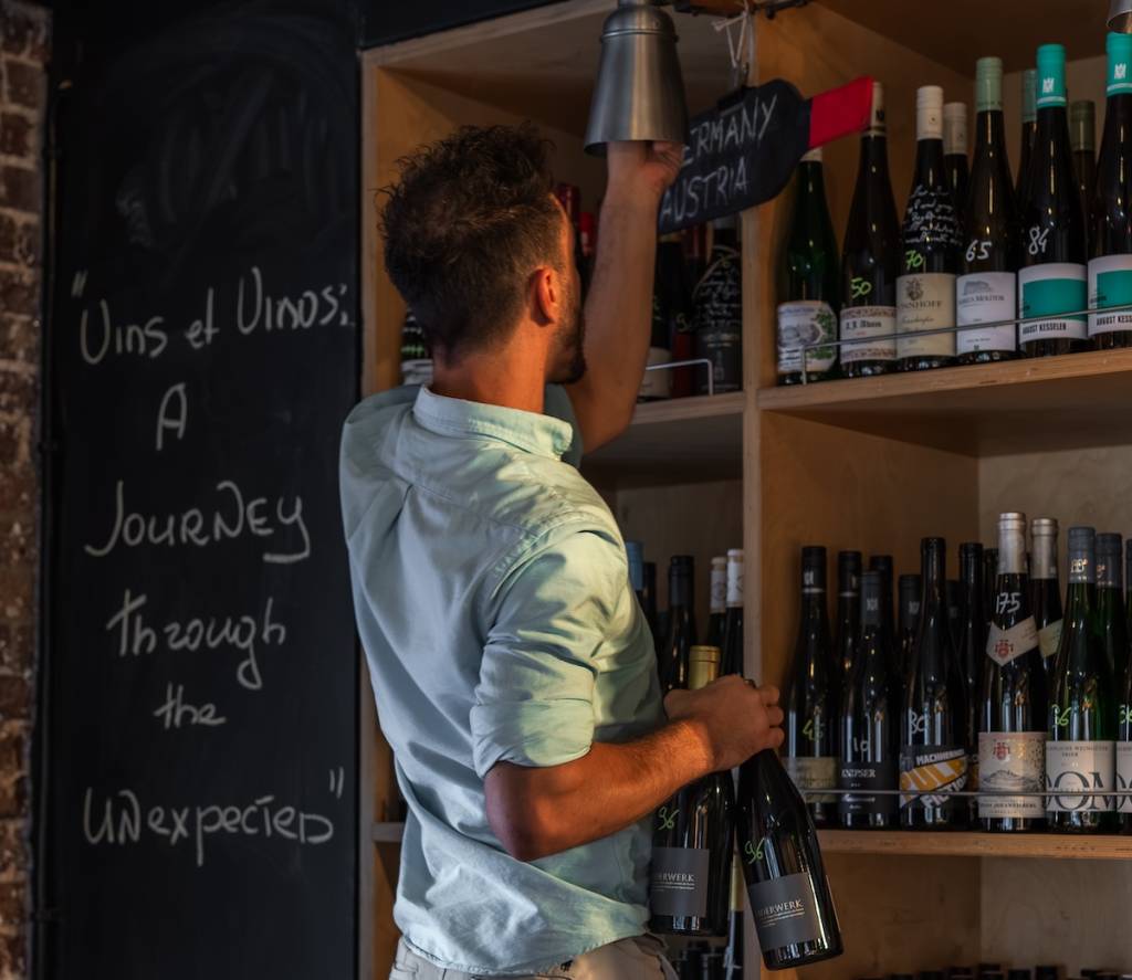 A man ringing a bell in front of a shelf of wines, with a chalkboard reading 'A journey through the Unexpcted'