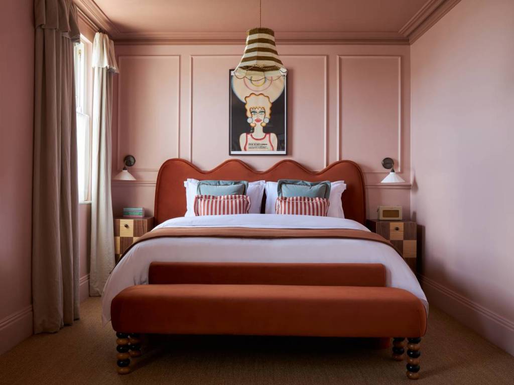 A bedroom with pink walls, a quirky painting above the bed, a large bed with a dark terracotta coloured headboard with white linen sheets.