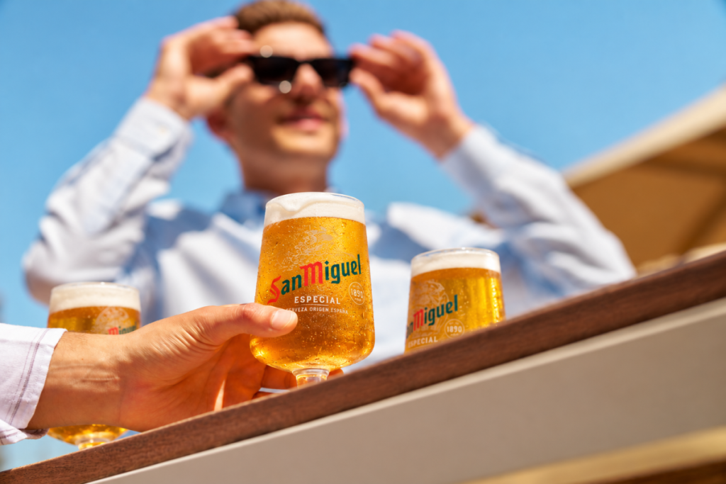 In the foreground a hand is holding a pint of San Miguel, in the background a smiling man is adjusting his sunglasses