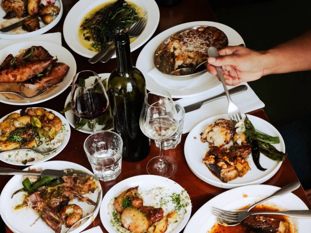 una tavola imbandita con piatti tipici del Sunday Roast, dalla carne alle patate e ai contorni, oltre a bottiglie e bicchieri di vino