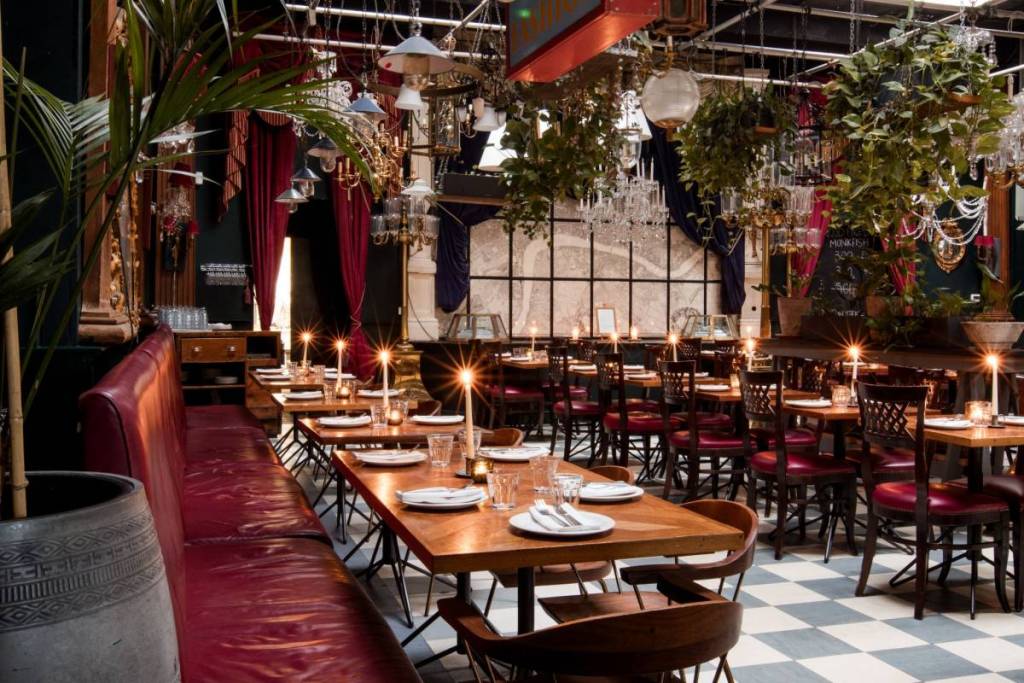 Rows of dining tables in a restaurant underneath chandeliers and plants
