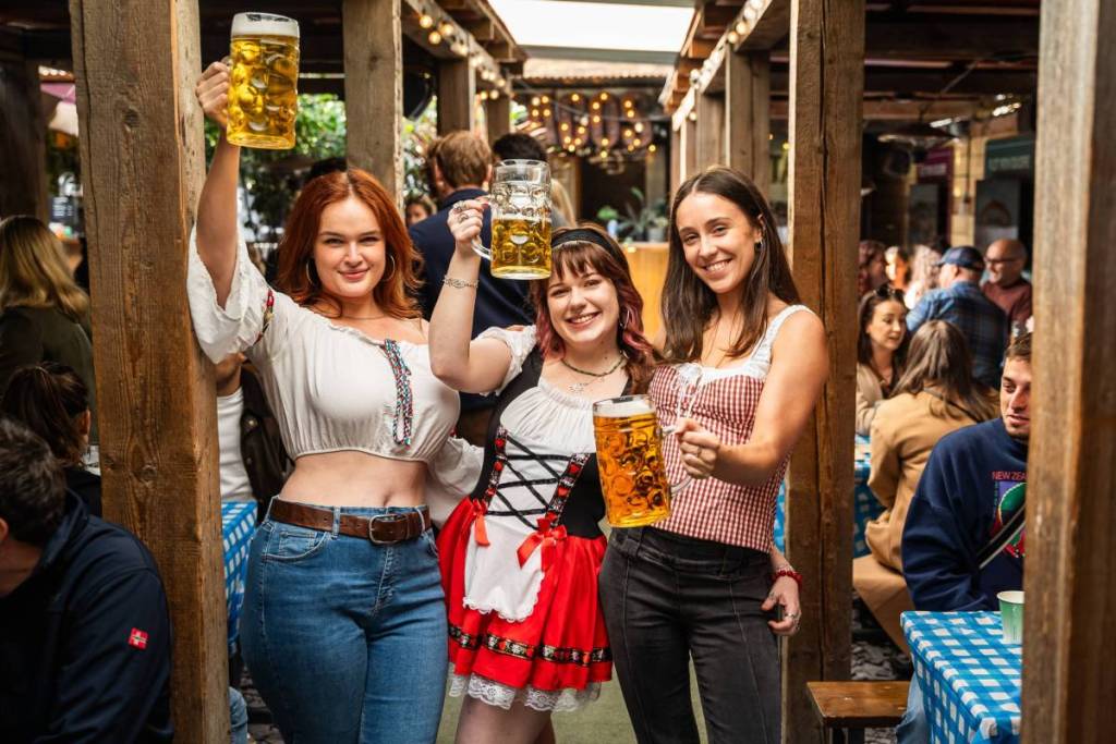 Drei Mädchen stehen auf einer Oktoberfest-Party, blicken in die Kamera und halten große Bierkrüge hoch