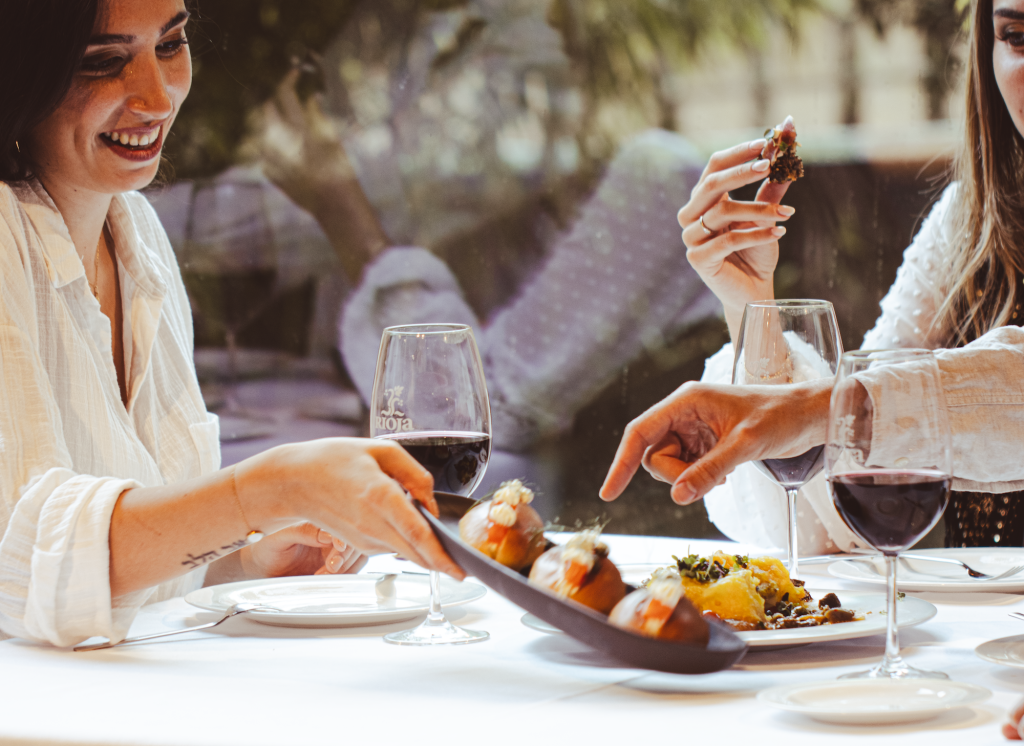 Two women at an outdoor restaurant table pick at tapas with glasses of wine