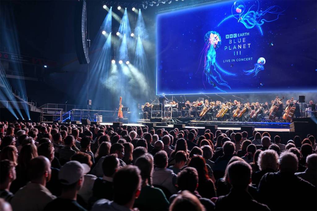 el público observa cómo una orquesta toca bajo una pantalla gigante en la que se ven medusas nadando alrededor de un título que reza: BBC Studios, Blue Planet III, Live In Concert