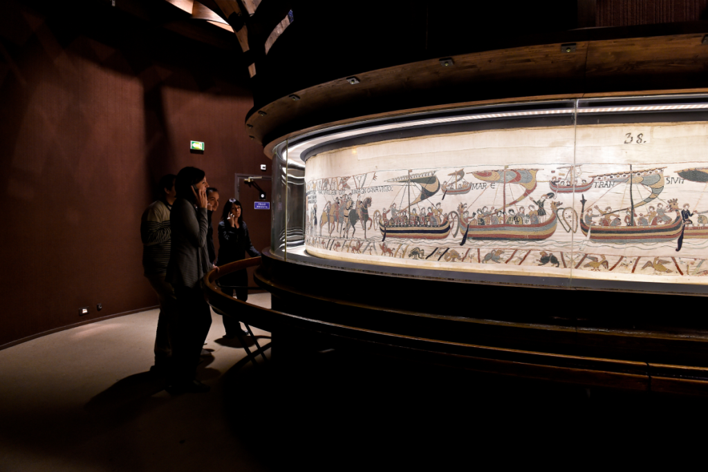 Pessoas a admirar a Tapeçaria de Bayeux — uma tapeçaria com bordados intricados, atualmente em exposição no Museu Britânico
