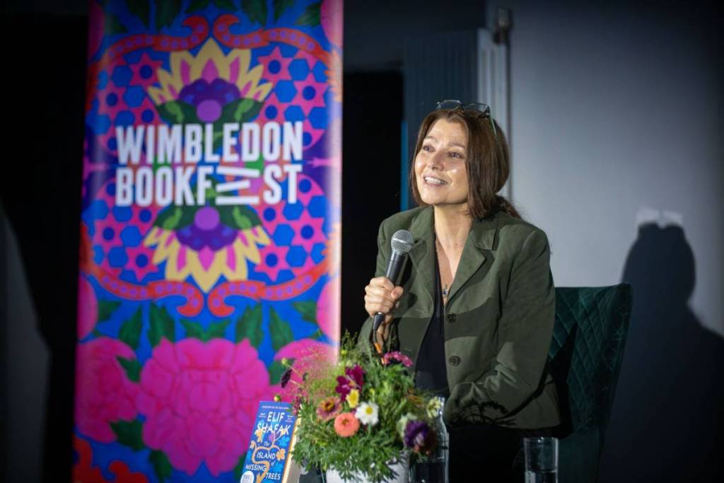 Elif Shafak holding a microphone, speaking to a crowd in front of a sign which reads 'Wimbledon Bookfest'